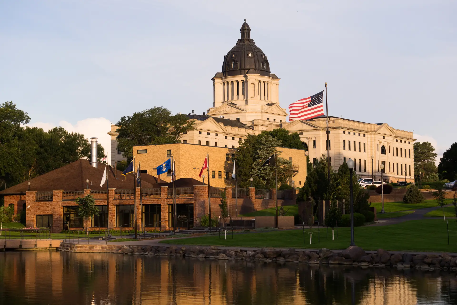 South Dakota state capitol building with American flag and surrounding greenery, reflecting in water.